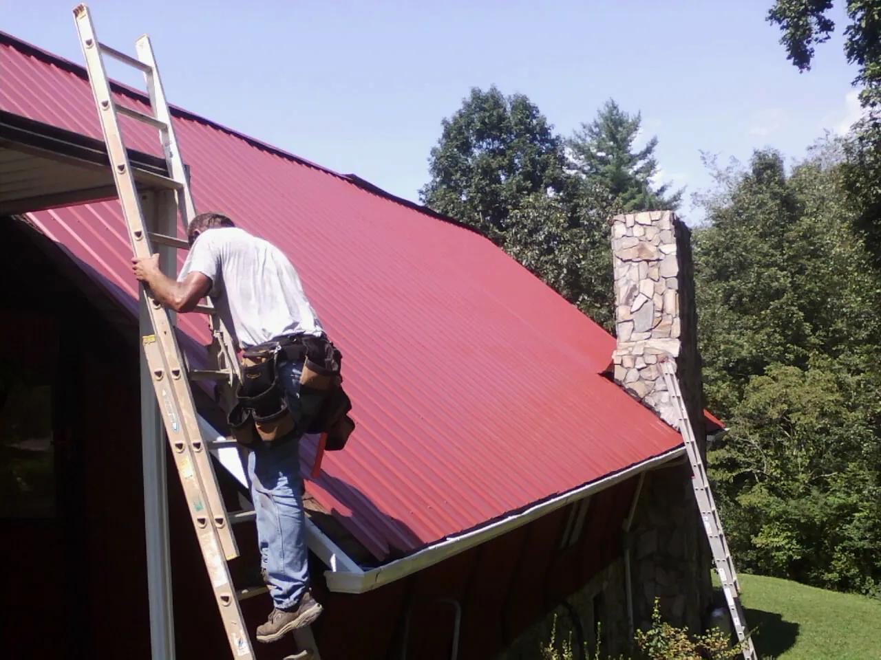Roofer on a ladder beside a red metal roof and stone chimney — Soffit & Fascia Repair by Roofing Brothers in Great Falls